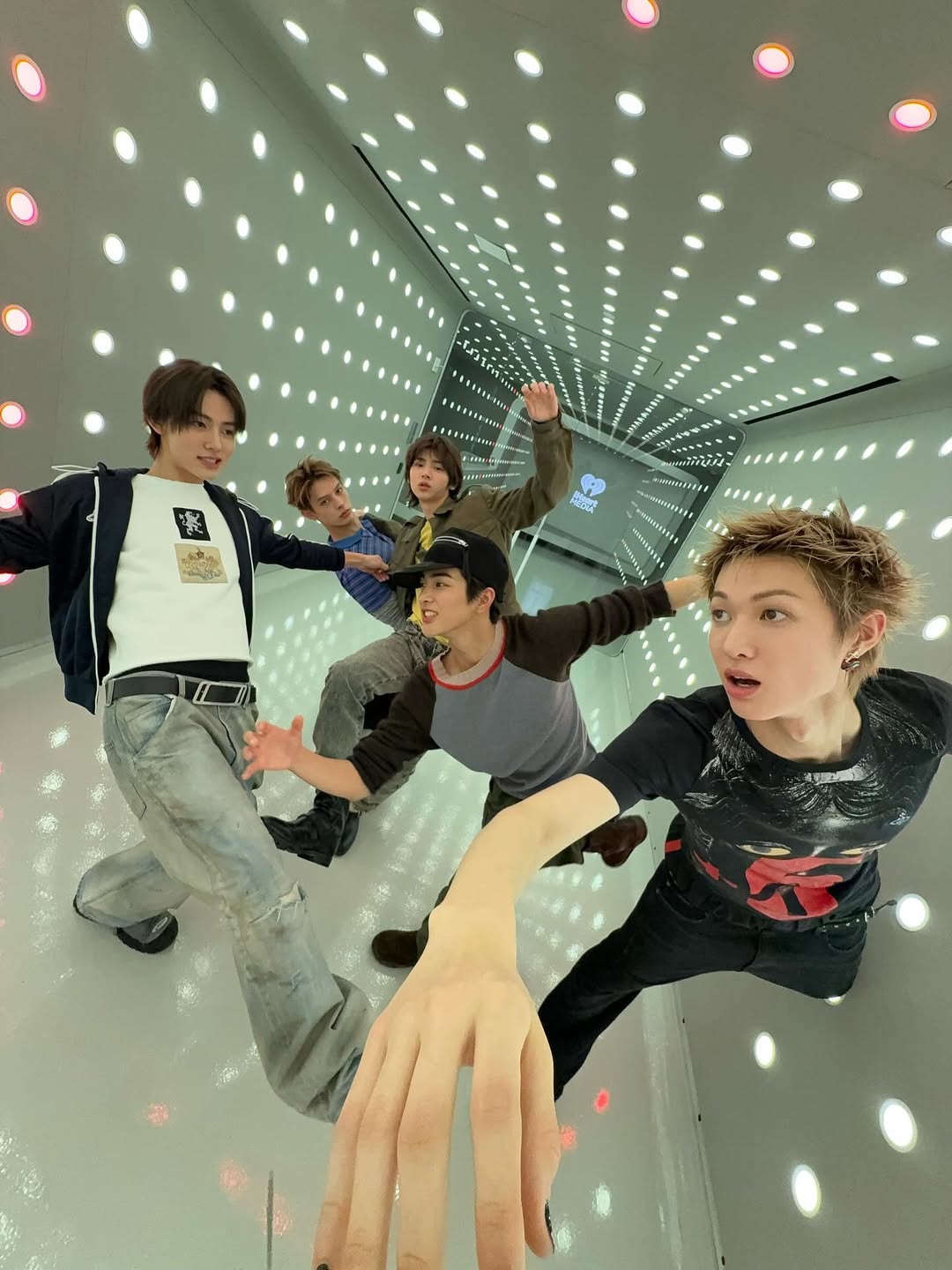 The five members of CORTIS in a mirrored room covered in lights, one hand reaching into the camera