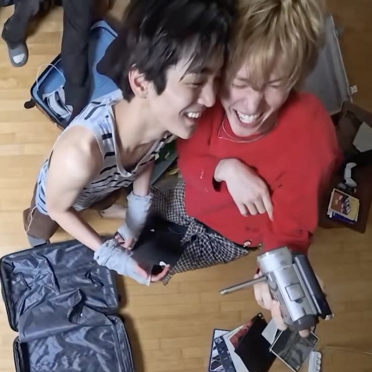 Martin and Keonho of CORTIS laughing together on the floor surrounded by open suitcases and a camera