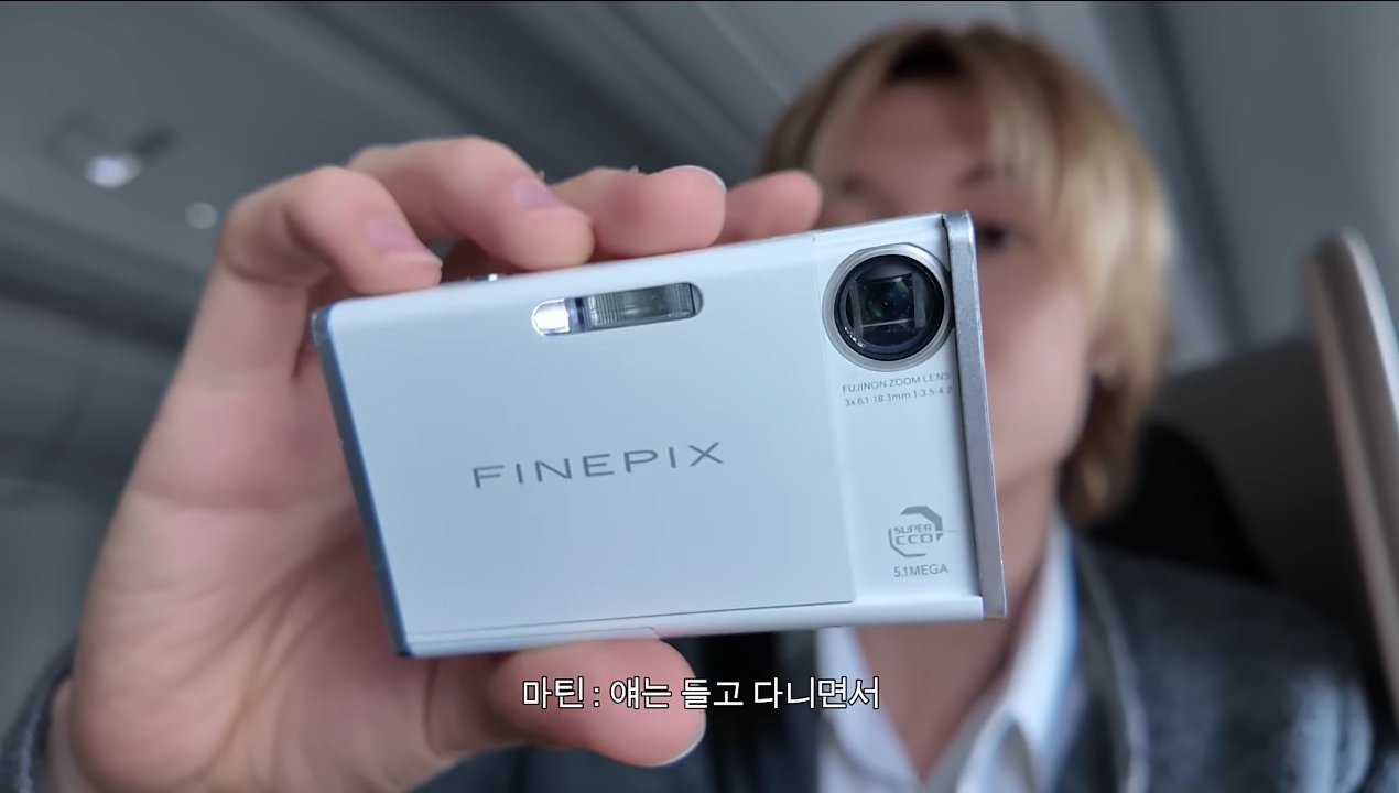 Martin of CORTIS holding up his white Fujifilm FinePix Z2 camera to the vlog camera