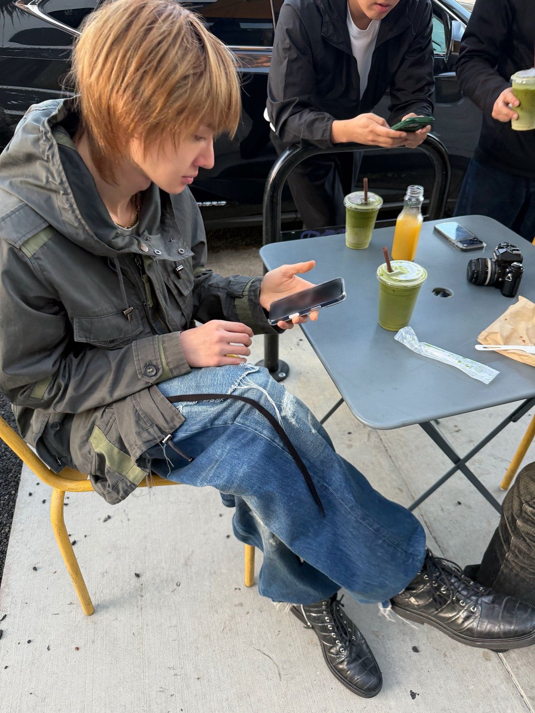 Martin of CORTIS sitting outside at a table with matcha drinks and a film camera, on his phone