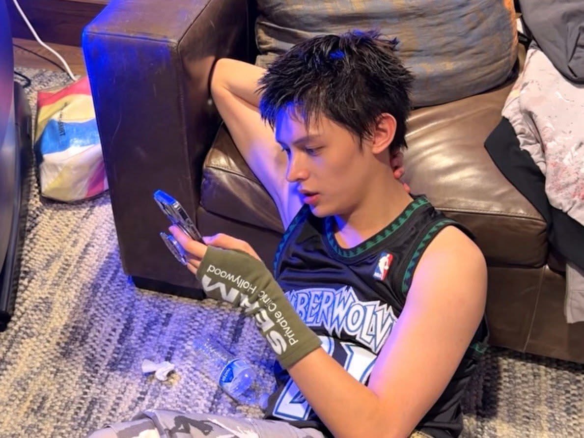 James of CORTIS sitting on the floor against a sofa in a Timberwolves jersey, scrolling on his phone