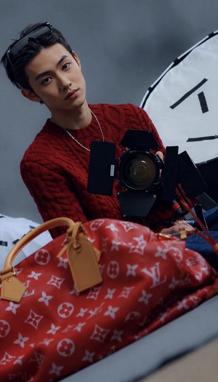 Seonghyeon of CORTIS in a red Louis Vuitton cable knit with sunglasses pushed up, behind a red monogram Speedy bag and a studio light