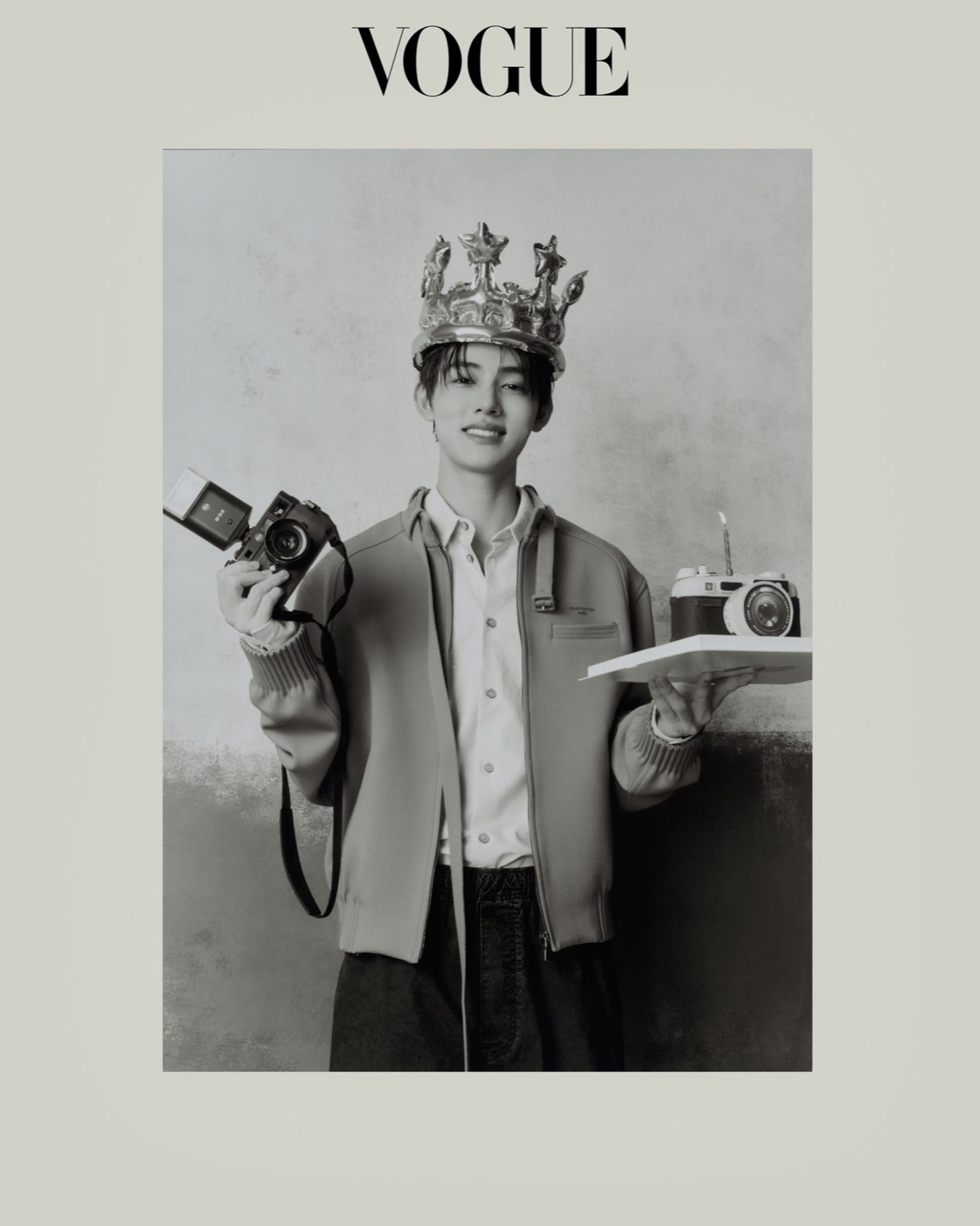 Seonghyeon of CORTIS in black and white, wearing a foil crown and holding a camera in one hand and a camera shaped cake in the other