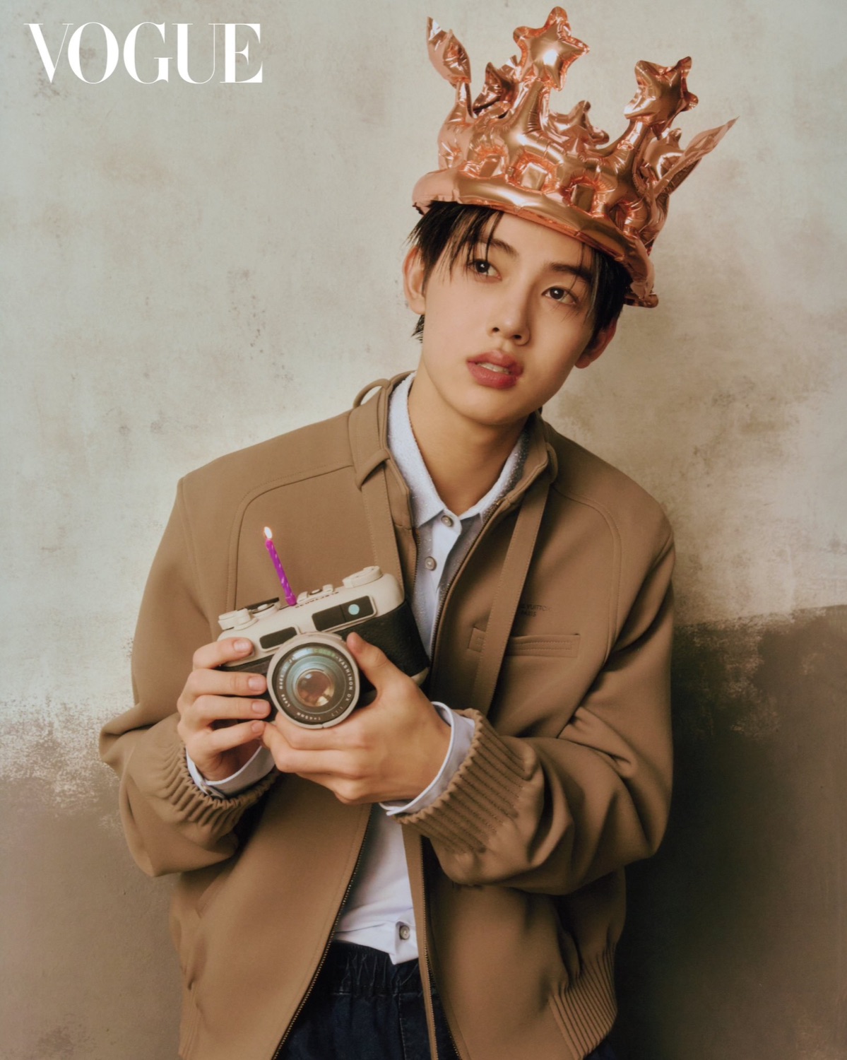 Seonghyeon of CORTIS in a beige Louis Vuitton jacket wearing a foil crown and holding a film camera with a birthday candle on it