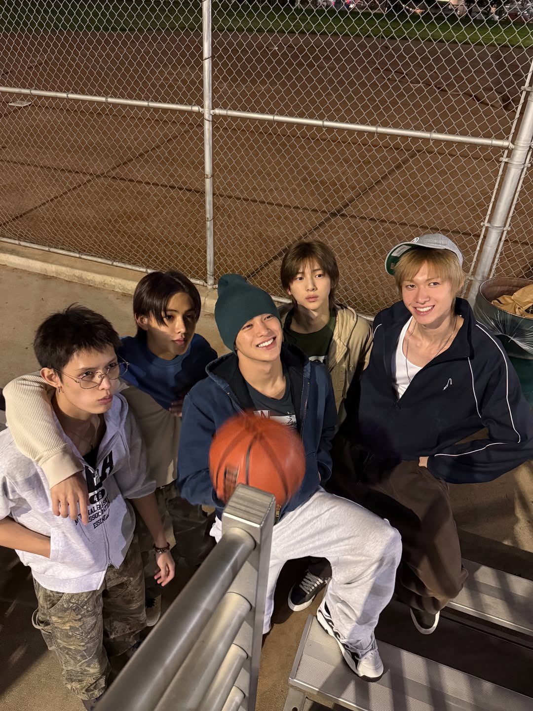 The five members of CORTIS sitting together on the bleachers of a basketball court at night